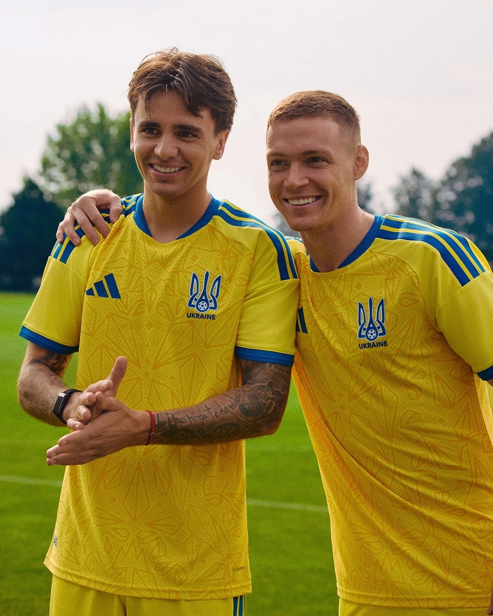 Двоє футболістів на фоні тренувального поля в жовто-блакитній формі збірної України, усміхаються перед камером.