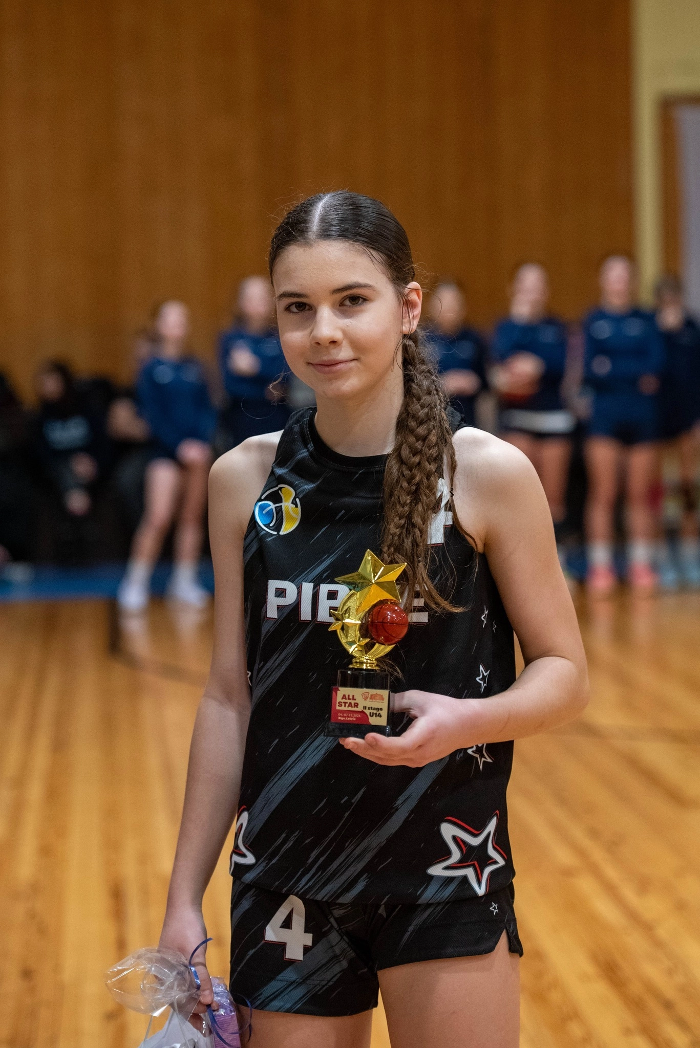 Юна баскетболістка в спортивній формі тримає трофей на змаганнях, інші гравці на задньому плані