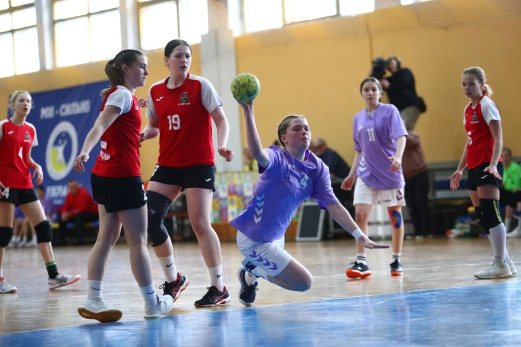 Юна гандболістка в фіолетовій формі виконує кидок у матчі проти команди в червоних майках