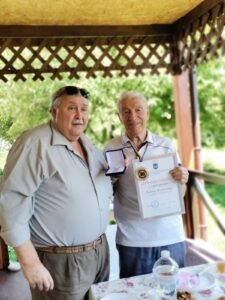 Двоє чоловіків на веранді, один тримає нагороду та грамоту, інший стоїть поруч