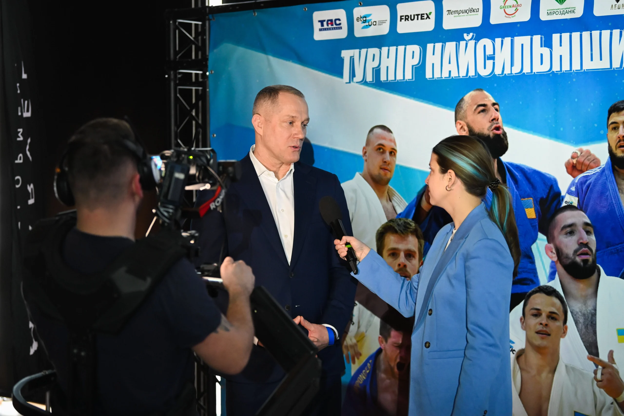 Чоловік в костюмі дає інтерв'ю журналістці на фоні банера Турніру найсильніших з дзюдо у Львові