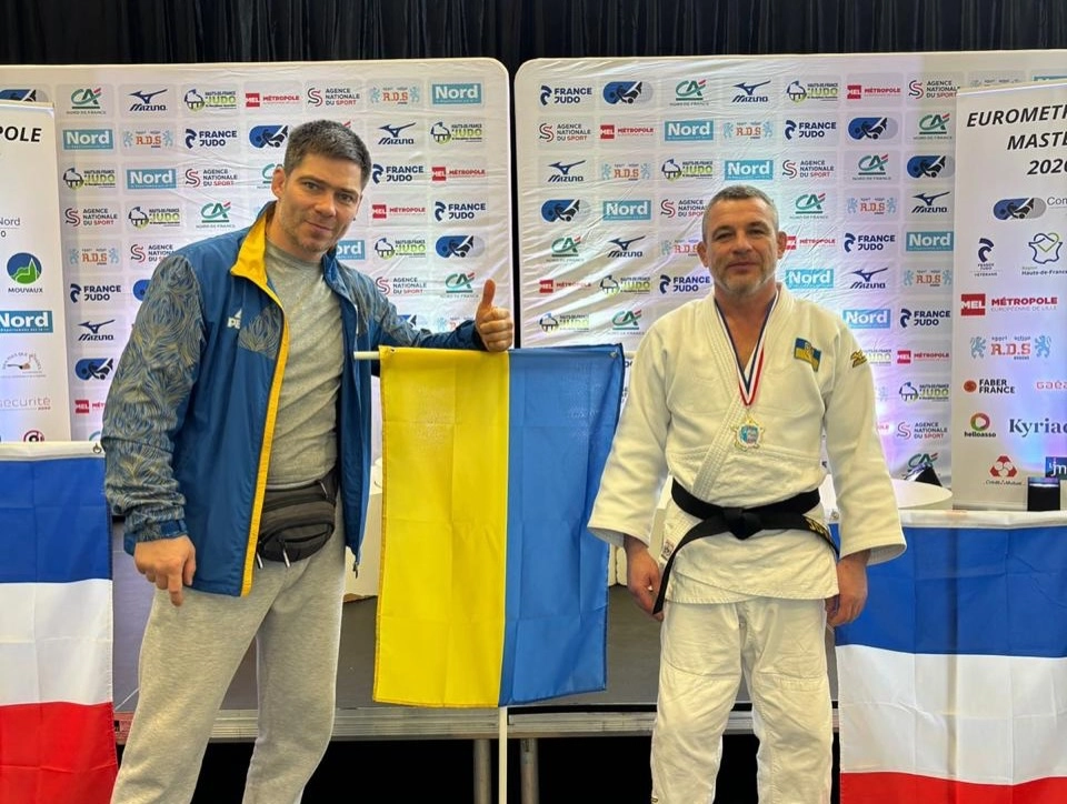 Двоє чоловіків у спортивному одязі стоять біля українського прапора на турнірі з дзюдо в Муво