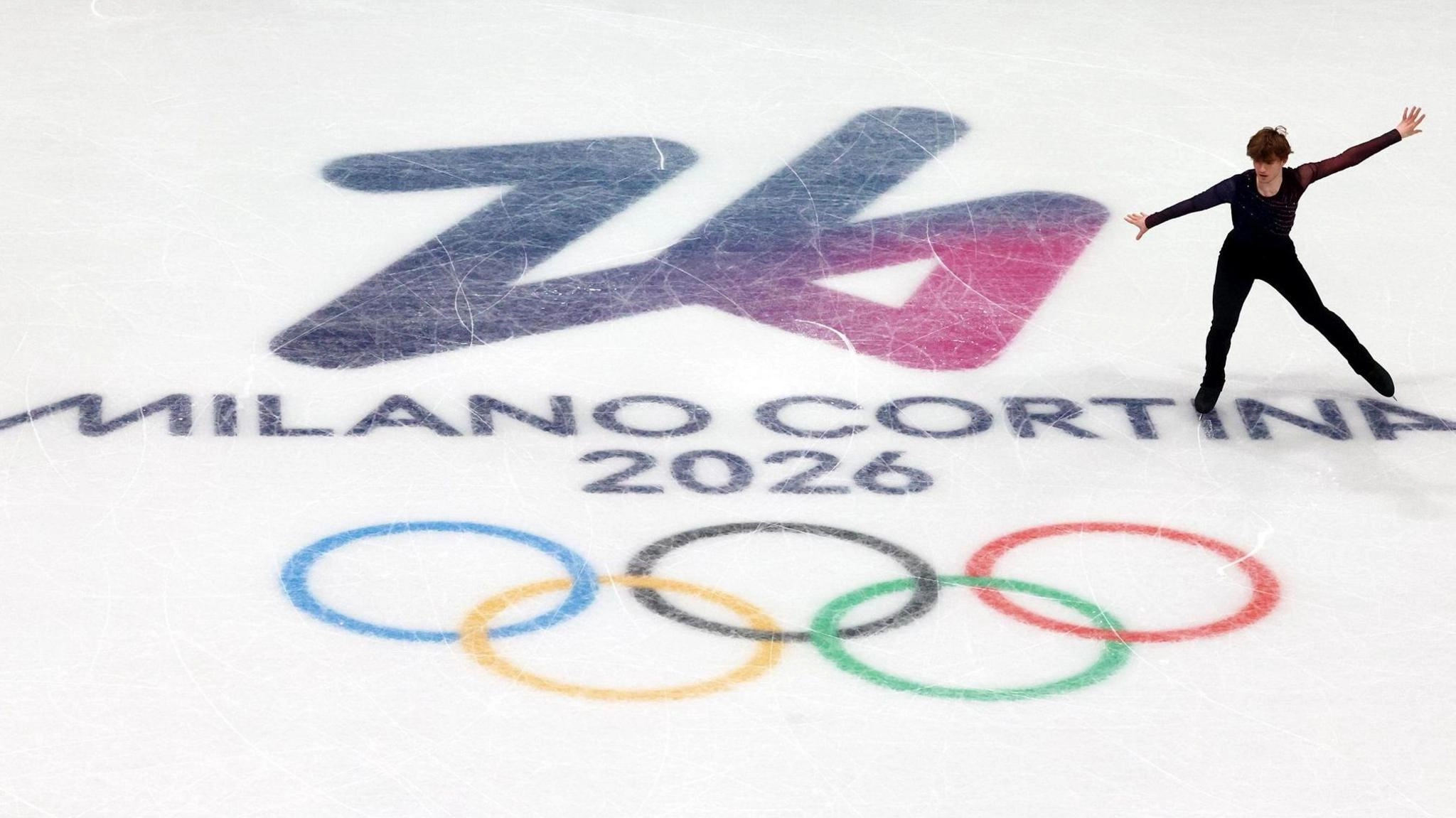 Фігурист виконує елемент на льоду з логотипом Мілан-Кортіна 2026 та олімпійськими кільцями