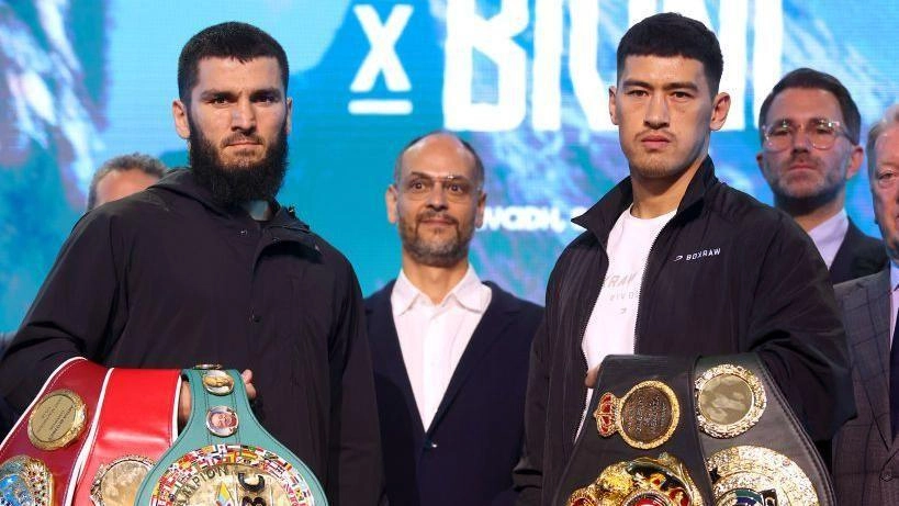 Боксери з чемпіонськими поясами стоять поруч на пресконференції перед боєм, позаду них офіційні особи