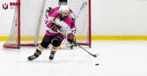 Хокеїстка в рожево-білій формі веде шайбу на льоду перед воротами під час матчу