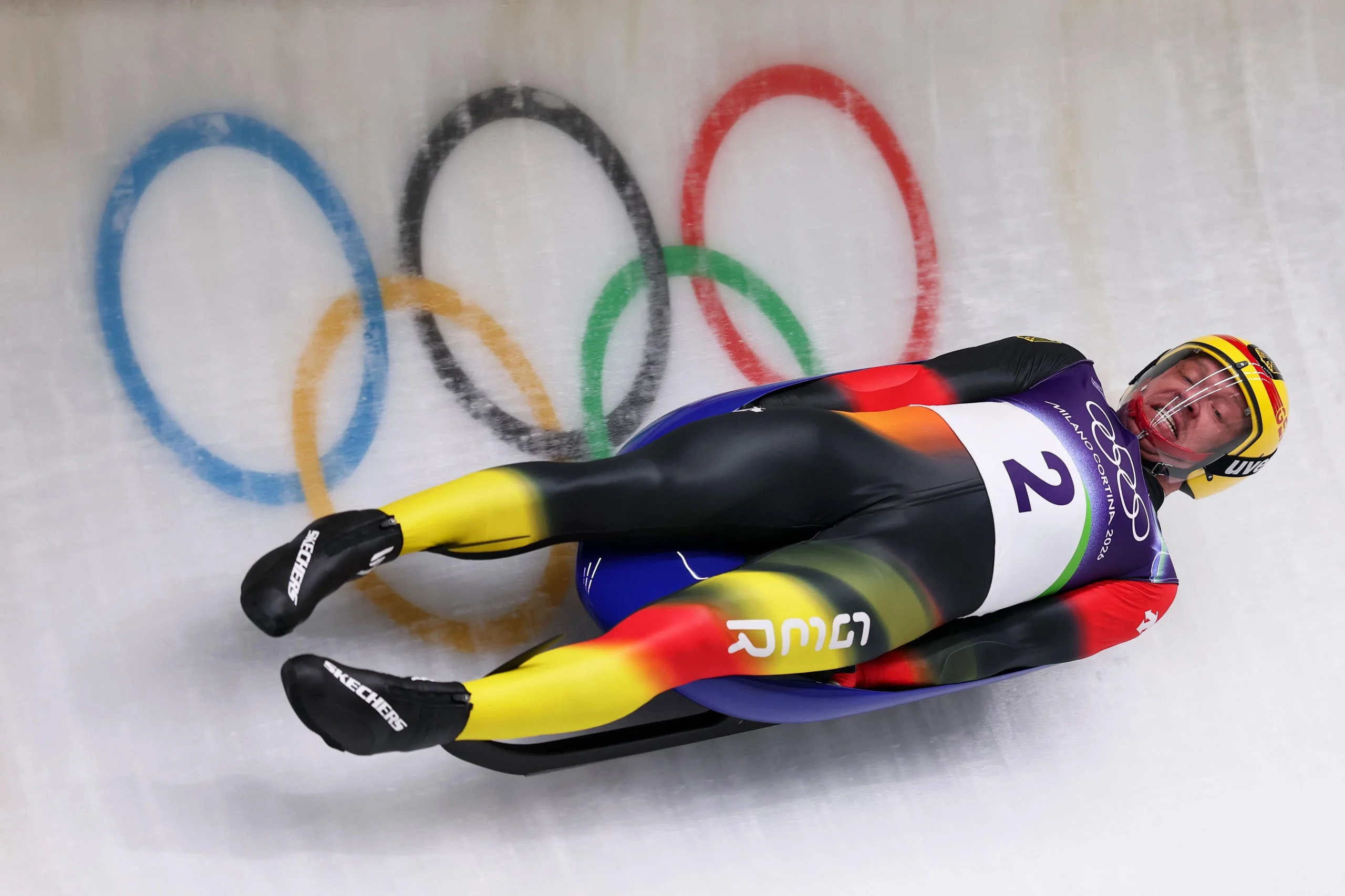 Спортсмен на санях змагається на трасі, на фоні олімпійських кілець, демонструючи швидкість і майстерність