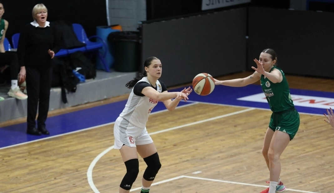 Баскетболістка в білій формі робить пас, захисниця в зеленій формі намагається перехопити м'яч