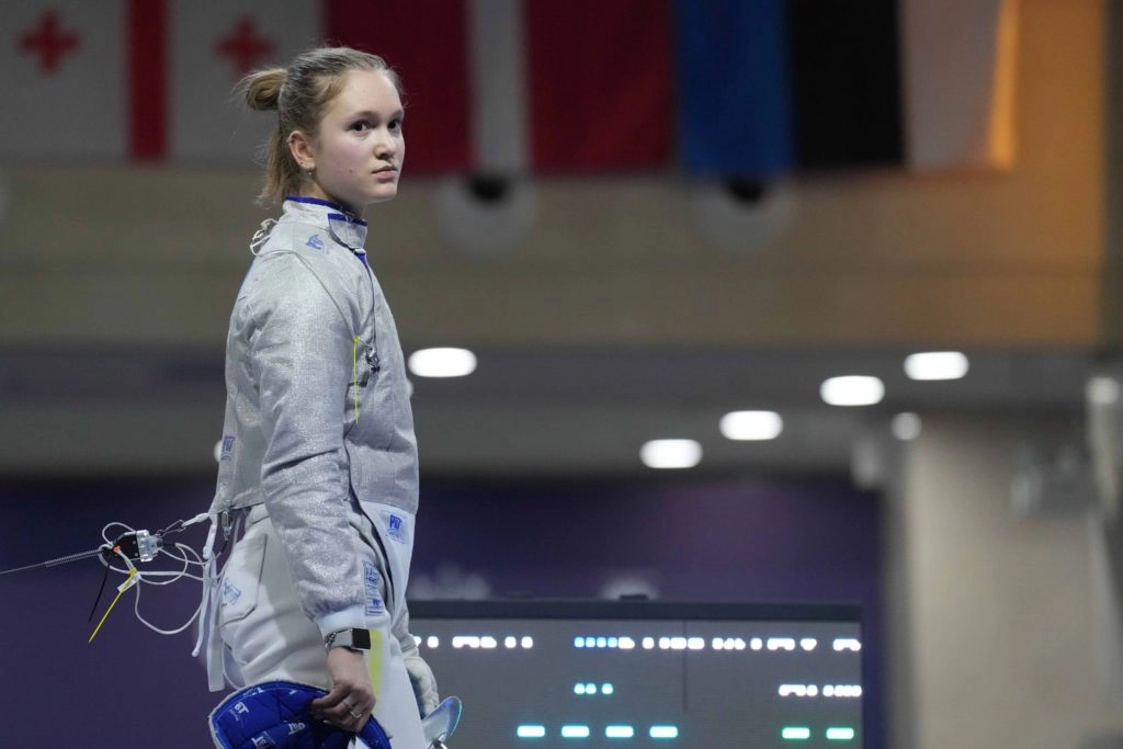 Шаблістка Майя Величко на чемпіонаті Європи, тримає шолом, на тлі прапорів Грузії, Латвії, Естонії