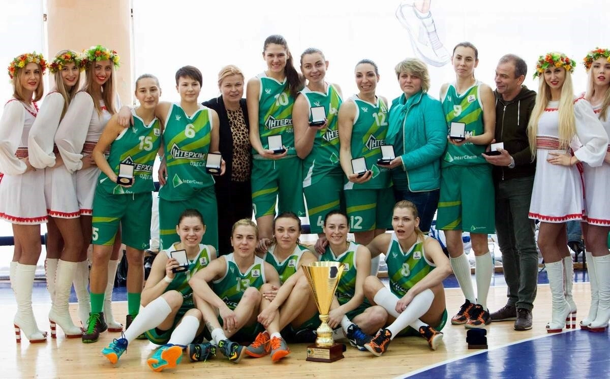 Жіноча баскетбольна команда з кубком на майданчику, оточена вболівальниками в національних костюмах