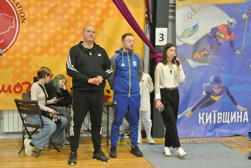 Троє людей стоять на сцені під час змагань з фехтування у Броварах, ведуча тримає мікрофон