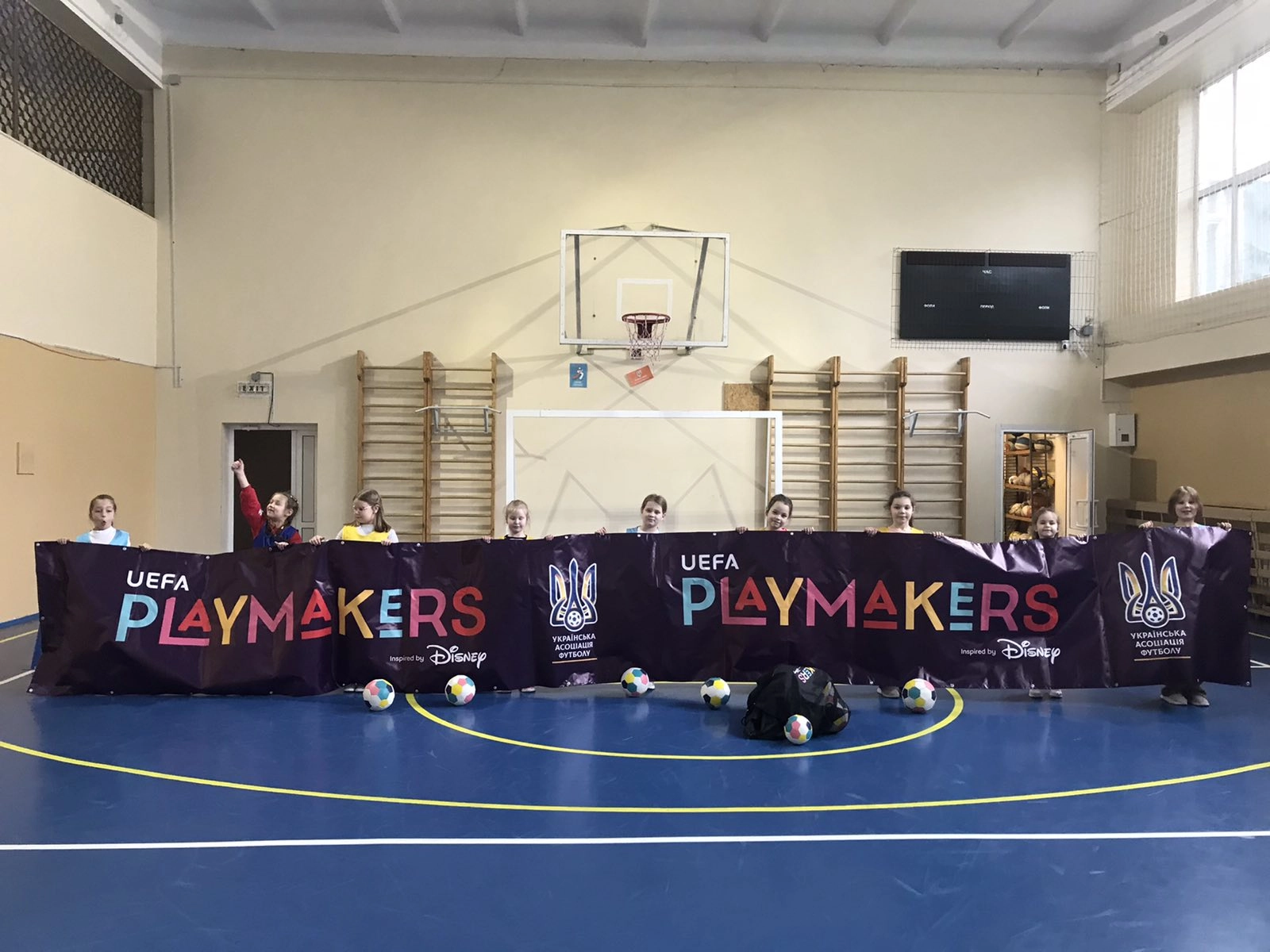 Дівчата тримають банер UEFA Playmakers у спортивному залі школи, перед ними футбольні м'ячі