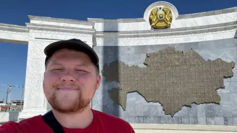 Чоловік у червоній футболці фотографується перед мапою Казахстану на стіні з гербом зверху