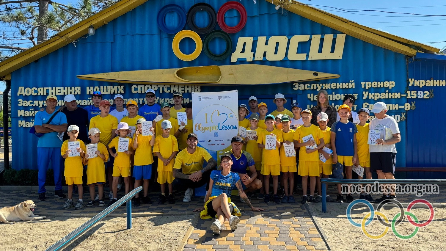 Група молодих спортсменів у жовтій формі з тренерами на фоні спортивного об'єкта з написом "ДЮСШ