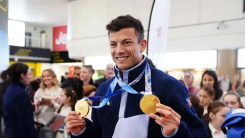 Олімпійський чемпіон з двома золотими медалями в аеропорту, оточений родиною та шанувальниками