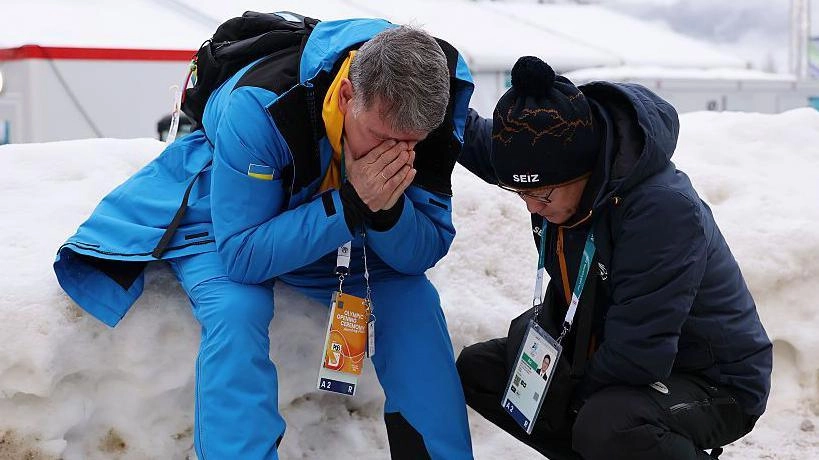 Український тренер у синій формі сидить на снігу, закривши обличчя руками, поруч стоїть інша людина