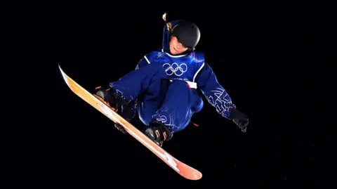 Сноубордистка виконує трюк у повітрі на зимових Олімпійських іграх, одягнена в синій костюм з емблемою Олімпіади