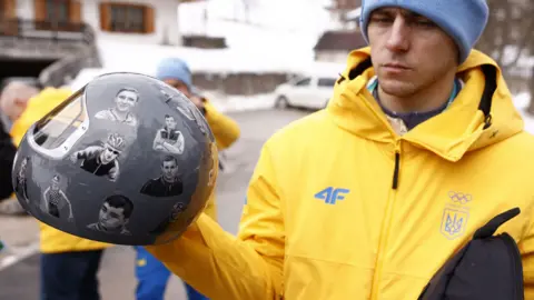 Український спортсмен тримає шолом з портретами загиблих атлетів на тлі зимового пейзажу