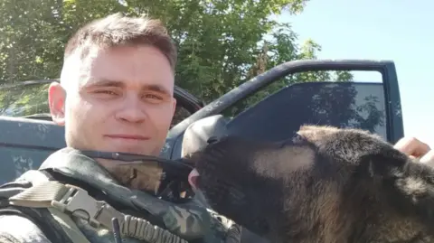 Український військовий в захисній формі посміхається поруч із собакою на фоні автомобіля та дерев