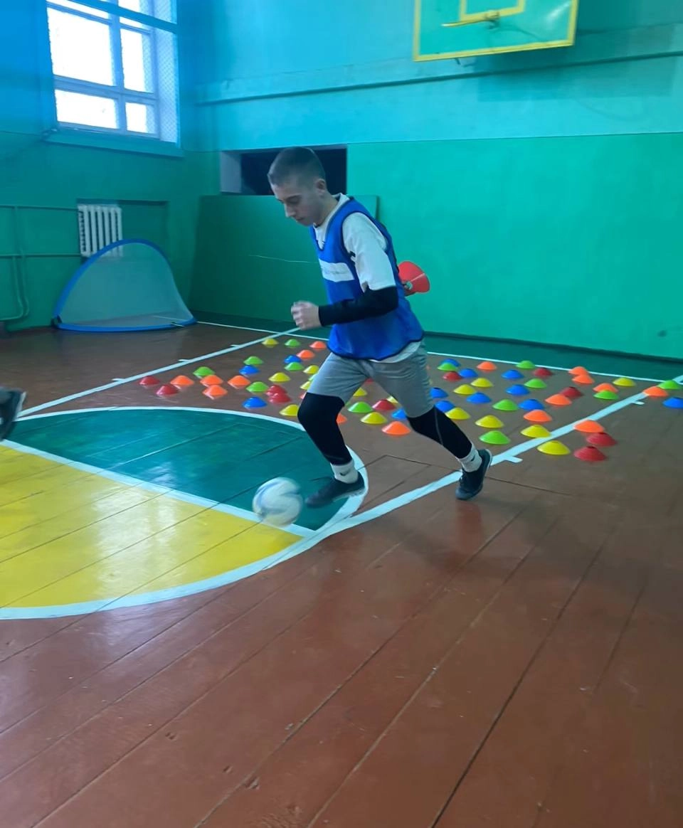 Юнак у спортивній формі веде м'яч у залі, на задньому плані розставлені кольорові конуси