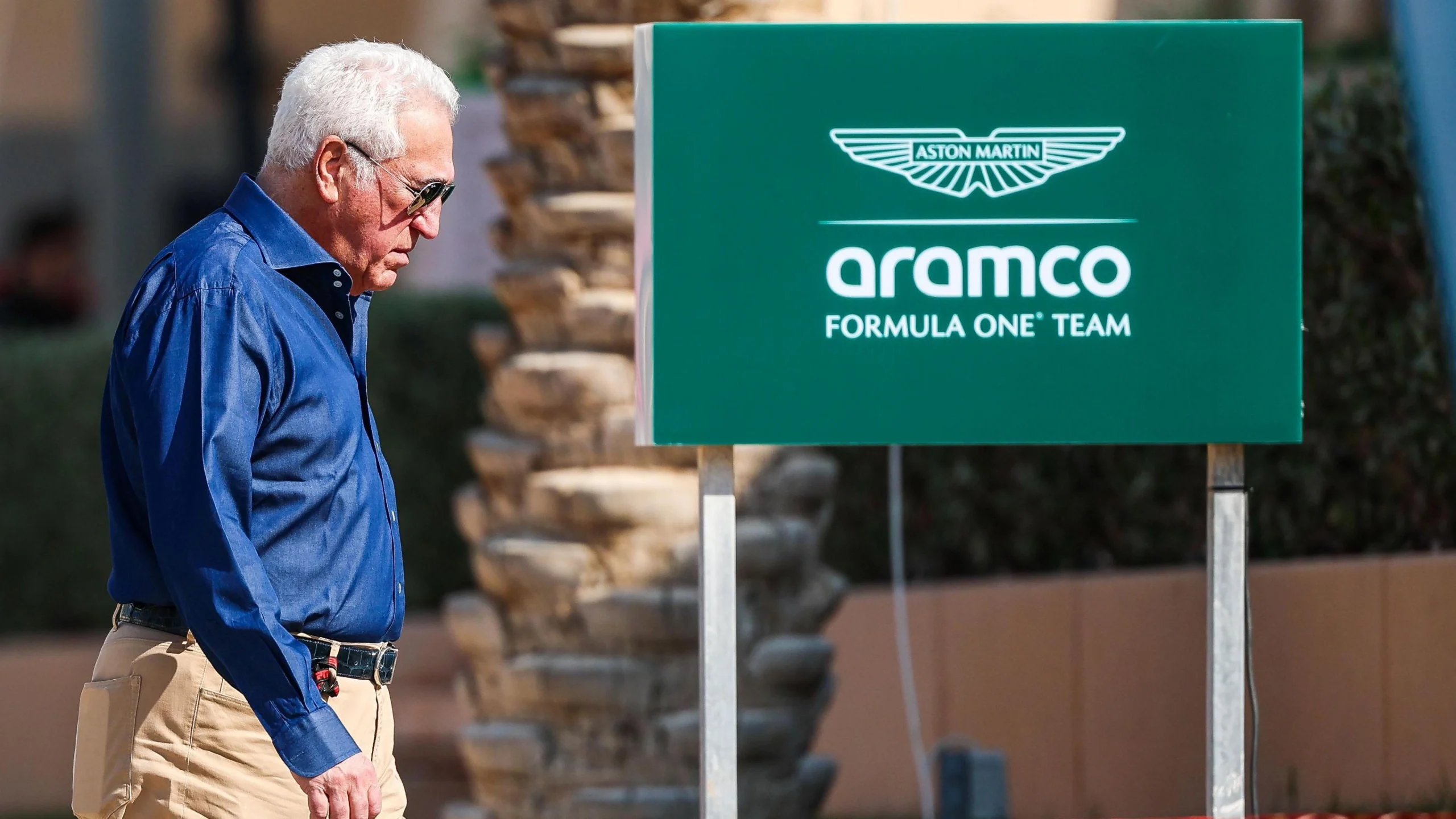 Лоуренс Стролл йде повз знак команди Aston Martin Aramco Formula 1 на тлі пальми