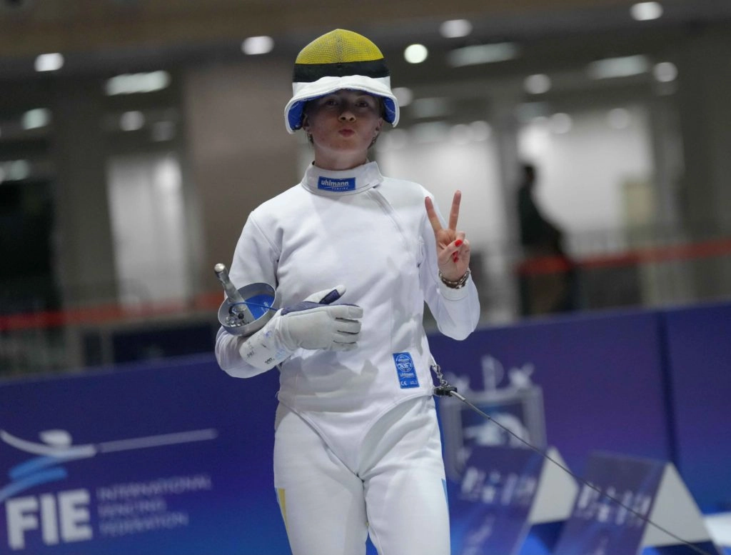 Українська шпажистка в білому костюмі на змаганнях, показує знак перемоги, тримаючи шпагу в руці