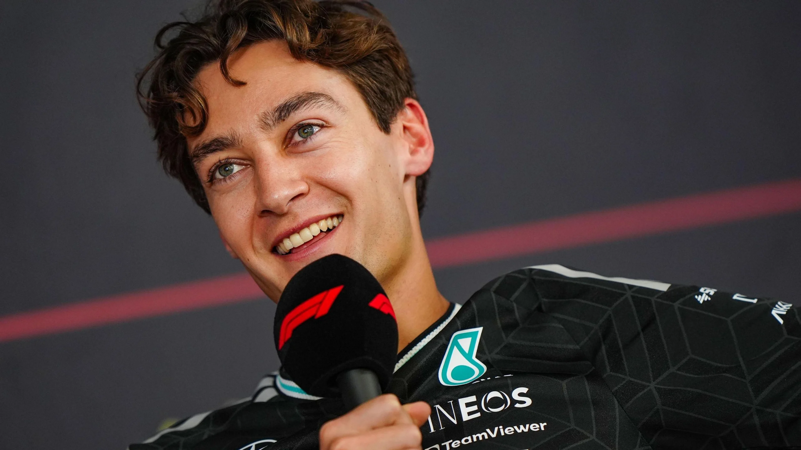 Молодий чоловік у формі Mercedes тримає мікрофон з логотипом F1, усміхаючись на прес-конференції