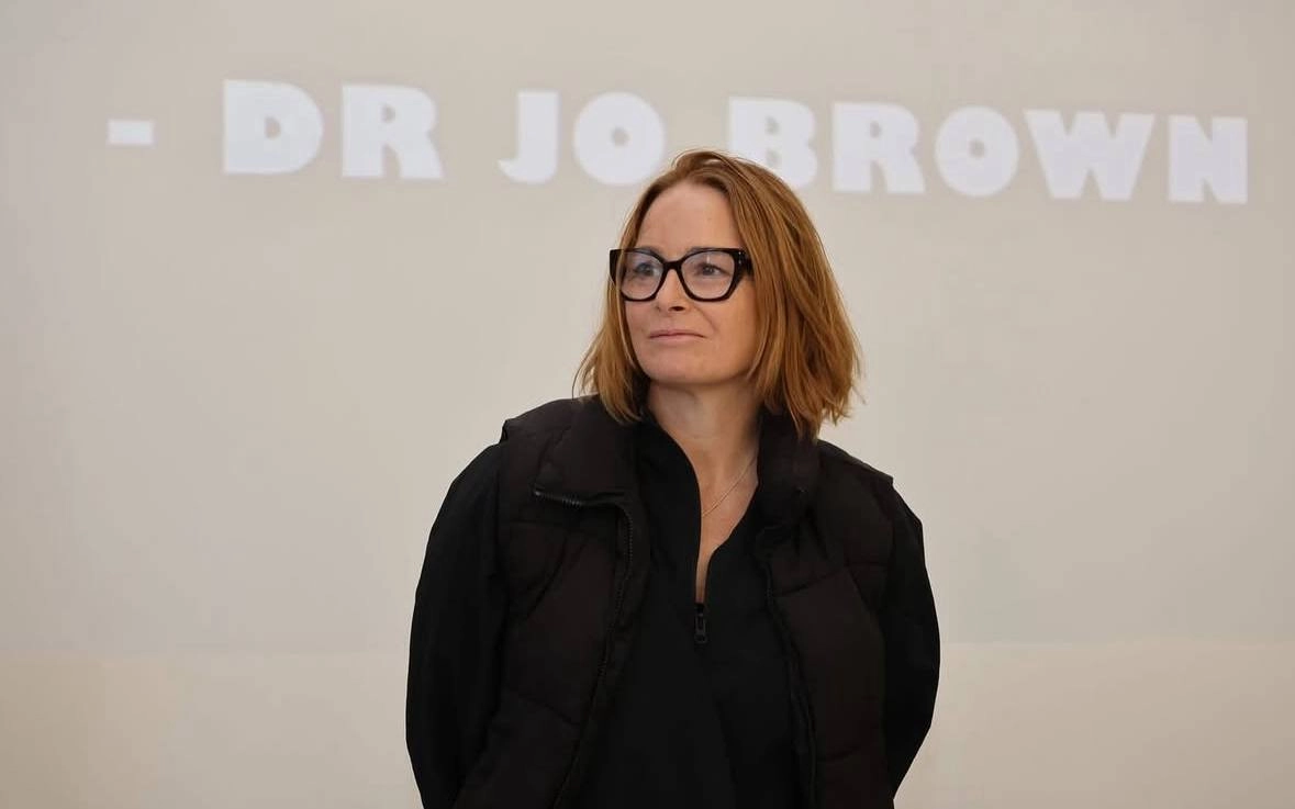 Жінка в окулярах стоїть перед екраном з написом "DR JO BROWN" під час лекції в Луцьку
