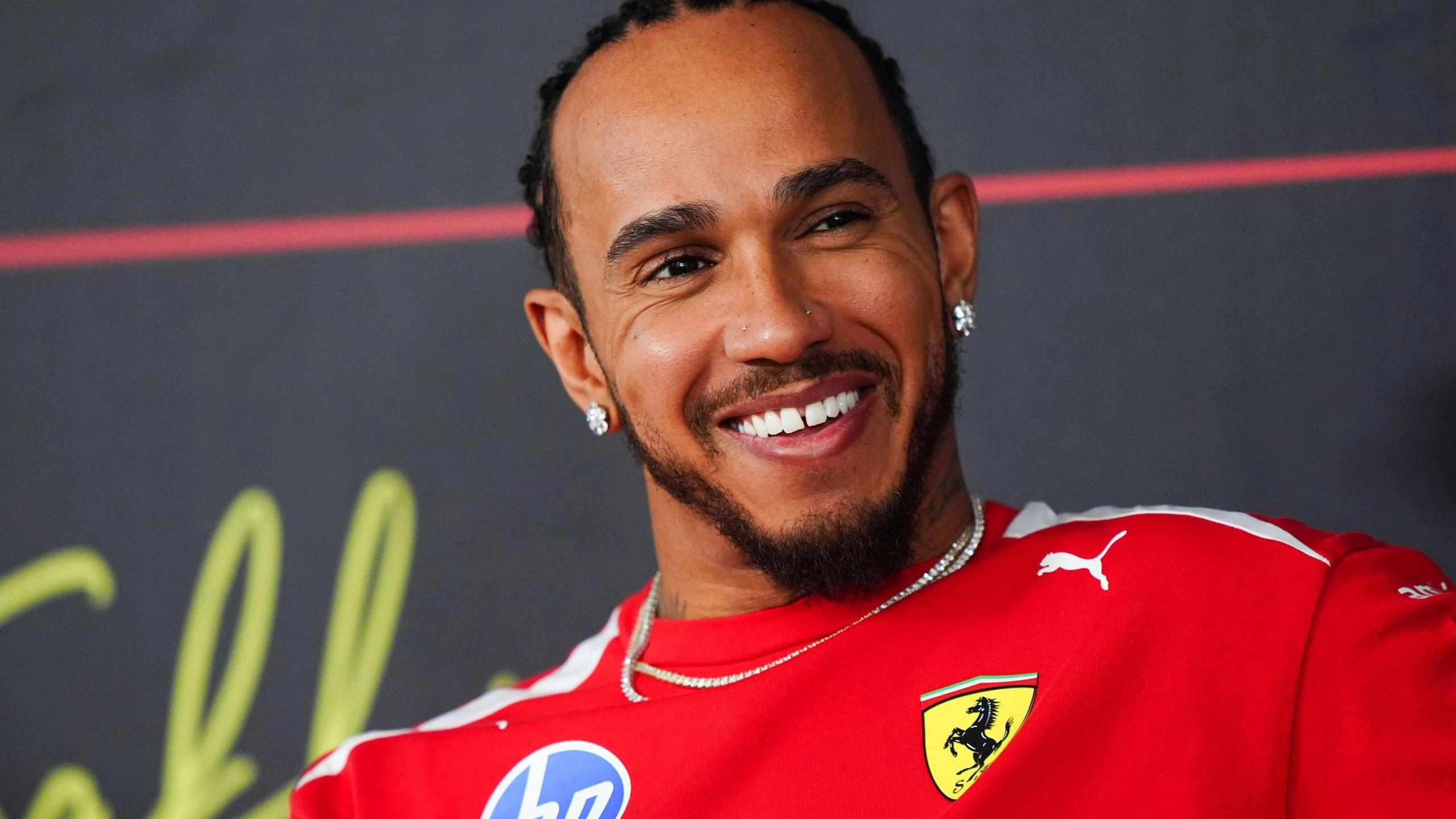 Чоловік у червоній формі Ferrari з усмішкою на обличчі на фоні темної стіни з логотипами