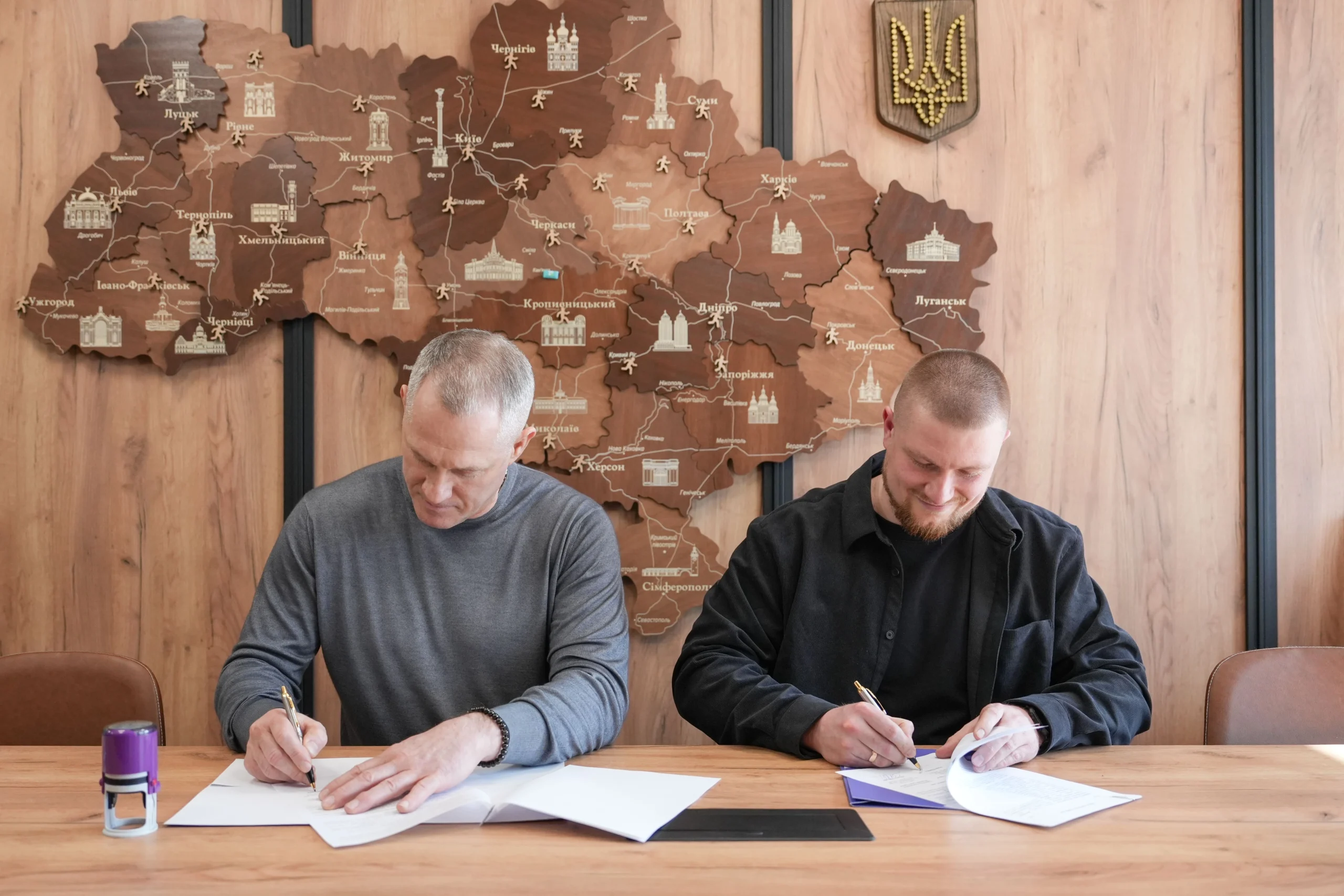 Двоє чоловіків підписують документи на фоні карти України та герба в офісі