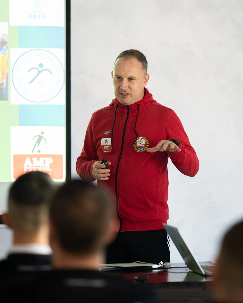Чоловік у червоній кофті проводить лекцію про ампфутбол перед групою слухачів на тлі презентації