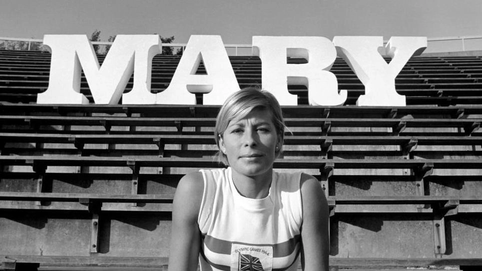 Жінка-спортсменка сидить на трибунах стадіону перед великими літерами "MARY" на задньому плані
