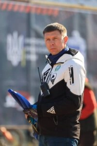 Чоловік у спортивній куртці тримає рацію на фоні спортивного заходу, зосереджений на події