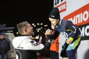 Чоловік вітає юного мотогонщика на подіумі під час змагань, нічне освітлення, радісна атмосфера